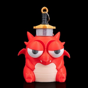 May include: A red dragon figurine with a sword on its head. The dragon has a grumpy expression with white and black eyes, small wings, and a light tan belly. The sword has a gold hilt and a silver blade. The background is black.