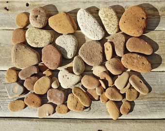 May include: A collection of various-sized, smooth, and textured stones in shades of tan, brown, and beige. The stones are arranged on a weathered wooden surface, with natural sunlight casting shadows. The assortment includes rounded, oval, and irregular shapes.