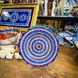 Peut inclure: Une assiette en céramique décorative avec un motif concentrique bleu, rouge et blanc détaillé. L'assiette est présentée sur un support transparent, mettant en valeur son design complexe. D'autres objets en céramique sont visibles en arrière-plan.