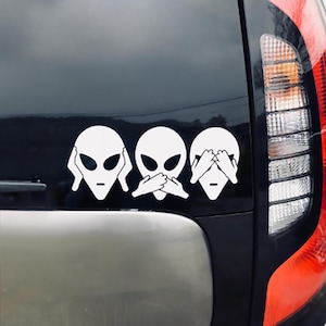 May include: Three white alien head stickers on a black car. The aliens are covering their eyes, ears, and mouth with their hands. The stickers are part of the "See no evil, hear no evil, speak no evil" saying.