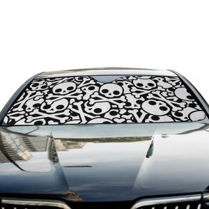 May include: A car windshield sun shade featuring a repeating black and white pattern of skulls and crossbones. The sun shade is positioned inside the front windshield of a dark-colored vehicle, covering the glass to block sunlight.