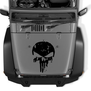 Könnte beinhalten: Ein schwarzer und weißer, abgenutzter Totenkopf-Aufkleber auf der Motorhaube eines grauen Jeep Wrangler.