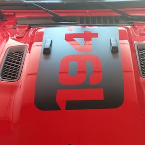 Könnte beinhalten: Rote Jeep Wrangler Motorhaube mit einem schwarzen und roten "1947" Aufkleber. Der Aufkleber ist mittig auf der Motorhaube angebracht und von zwei schwarzen Motorhaubenverschlüssen umgeben.