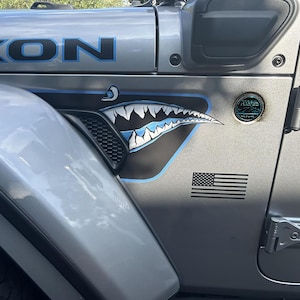 Può includere: Un pannello laterale di un veicolo color argento con una decalcomania a forma di bocca di squalo con denti bianchi e accenti blu. Il pannello include una bandiera americana nera, un badge circolare Trail Rated 4x4 e una decalcomania blu parziale.