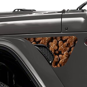 Può includere: Una Jeep Wrangler grigia con un adesivo a tema gingerbread man sul parafango. L'adesivo presenta un primo piano di biscotti gingerbread man.