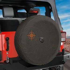 May include: A dark grey spare tyre cover featuring a thin orange topographic map pattern and a central compass rose design. The cover is mounted on the rear of a red vehicle, with a small camera lens visible in the centre of the tyre cover.