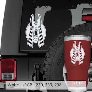 Puede incluir: Calcomanía blanca de la cabeza de un chacal Anubis en un coche negro con una luz trasera roja. La calcomanía también está en un vaso YETI rojo.