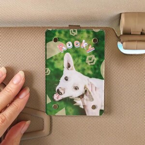 Peut inclure: Accessoire rectangulaire pour pare-soleil de voiture avec un chien blanc et le nom "Rocky" en texte coloré. Le fond présente un motif vert et géométrique. Fixé à un pare-soleil de voiture beige.