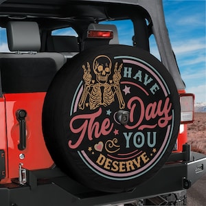 May include: A black spare tyre cover featuring a graphic of a skeleton making peace signs with its hands. The design includes the text "HAVE The Day YOU DESERVE" in pink and light blue, surrounded by small stars and hearts on a circular background.