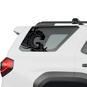 May include: A black vinyl decal featuring a stylised Japanese-inspired ocean wave design applied to the rear side window of a white sport utility vehicle. The graphic depicts swirling water currents and foam droplets in a bold, solid black colour.