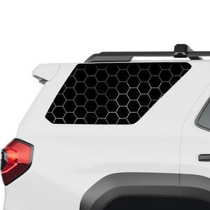 May include: A white vehicle rear quarter window featuring a black honeycomb pattern decal. The graphic consists of repeating black hexagons outlined in thin white lines, covering the entire surface of the rear side window of the car.