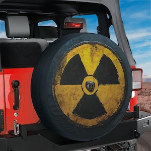 May include: A circular spare tire cover mounted on the back of a red vehicle. The cover features a distressed yellow and black radiation hazard symbol with a weathered texture. A small black camera lens is centered on the tire cover surface.