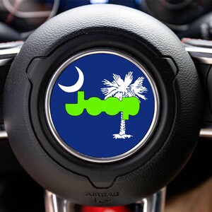 May include: A circular steering wheel emblem featuring a dark blue background with a bright lime green Jeep logo, a white crescent moon, and a white palmetto tree silhouette centered on the horn cover of a black leather steering wheel.