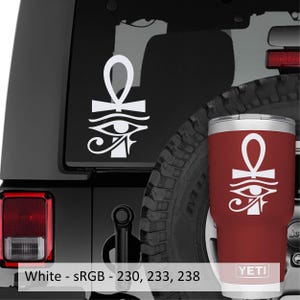 May include: A dark red stainless steel tumbler featuring a white graphic of an Egyptian ankh symbol positioned above the Eye of Horus. The brand name YETI is printed in white at the base of the tumbler, which matches the white decal on the vehicle.