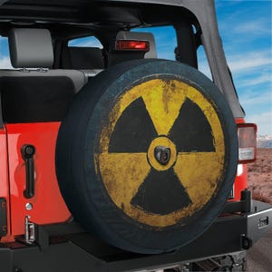 May include: A circular spare tire cover mounted on the back of a red vehicle. The cover features a distressed yellow and black radiation hazard symbol printed on the front, with a small camera lens centered in the middle of the design.
