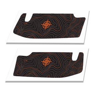 May include: Two identical black adhesive decals featuring a topographic map pattern with thin orange contour lines. Each decal displays a centred orange compass rose graphic with cardinal direction letters N, S, E, and W clearly visible.