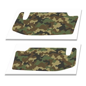 May include: Two identical automotive sun visor covers featuring a camouflage pattern in shades of forest green, olive, brown, and black. Each cover has a distinct contoured shape with a cutout section on one side, designed for vehicle interior use.