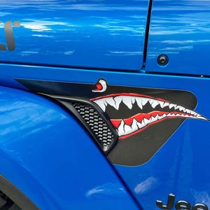 Può includere: Un pannello laterale di un veicolo blu brillante con una copertura nera per la presa d'aria decorata con una decalcomania grafica di una bocca di squalo. Il design include denti bianchi affilati, gengive rosse e un occhio nero sulla superficie.