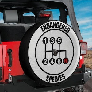 May include: A circular spare tire cover mounted on the back of a red vehicle. The cover is white with a black border and features a black manual transmission gear shift pattern with the text ENDANGERED SPECIES in black capital letters.