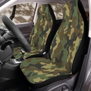 May include: Camouflage pattern car seat covers in shades of green, brown, and tan. The covers are designed to fit over the front seats of a vehicle. The steering wheel and dashboard are visible in the background.