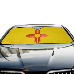 May include: A yellow car windshield sun shade featuring a red Zia sun symbol centered on the fabric. The rectangular shade is positioned inside the front windshield of a dark-colored vehicle, covering the glass to block sunlight.