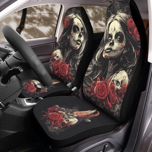 May include: Black car seat covers featuring a Day of the Dead design. The design includes a skull with floral accents and red roses. The seat covers are in a car interior with a steering wheel visible.