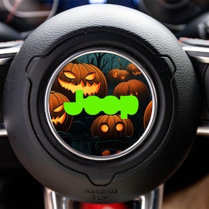 May include: Black steering wheel with a central emblem featuring a Halloween-themed design. The emblem showcases carved orange pumpkins against a dark background, with the word "Jeep" in bright green.