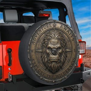 May include: A spare tire cover with a bronze-colored design featuring a compass and a Sasquatch face. The text "ADVENTURE AWAITS" and "EXPLORE THE WILDERNESS" are displayed around the edge. The cover is mounted on a red Jeep.
