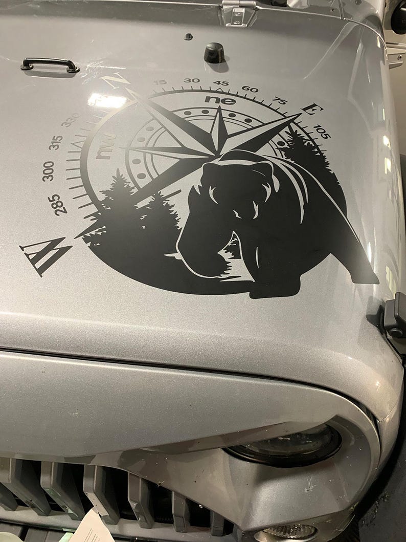 May include: A black and white compass decal with a bear silhouette and a compass rose. The compass has numbers from 15 to 105 in increments of 15. The compass is on a silver car hood.