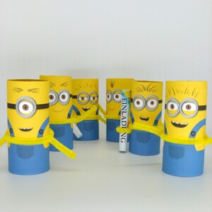 May include: Six yellow Minion characters made from cardboard toilet paper rolls. Each Minion has a blue jumpsuit, yellow goggles, and a black strap. The Minions are holding yellow pipe cleaners. One Minion is holding a white card with the word "Einladung" printed on it.