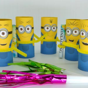 May include: Five yellow Minion character party favors made from cardboard toilet paper rolls. Each Minion has blue overalls, yellow hair, and googly eyes. The Minion in the center is holding a white card with the word "Einladung" printed on it.
