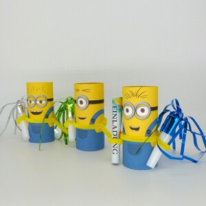May include: Three yellow and blue Minion-themed cardboard tubes with party favors inside. Each tube has a different party favor, including a green and white party blower, a blue and white party blower, and a small white box with the word "Einladung" printed on it.