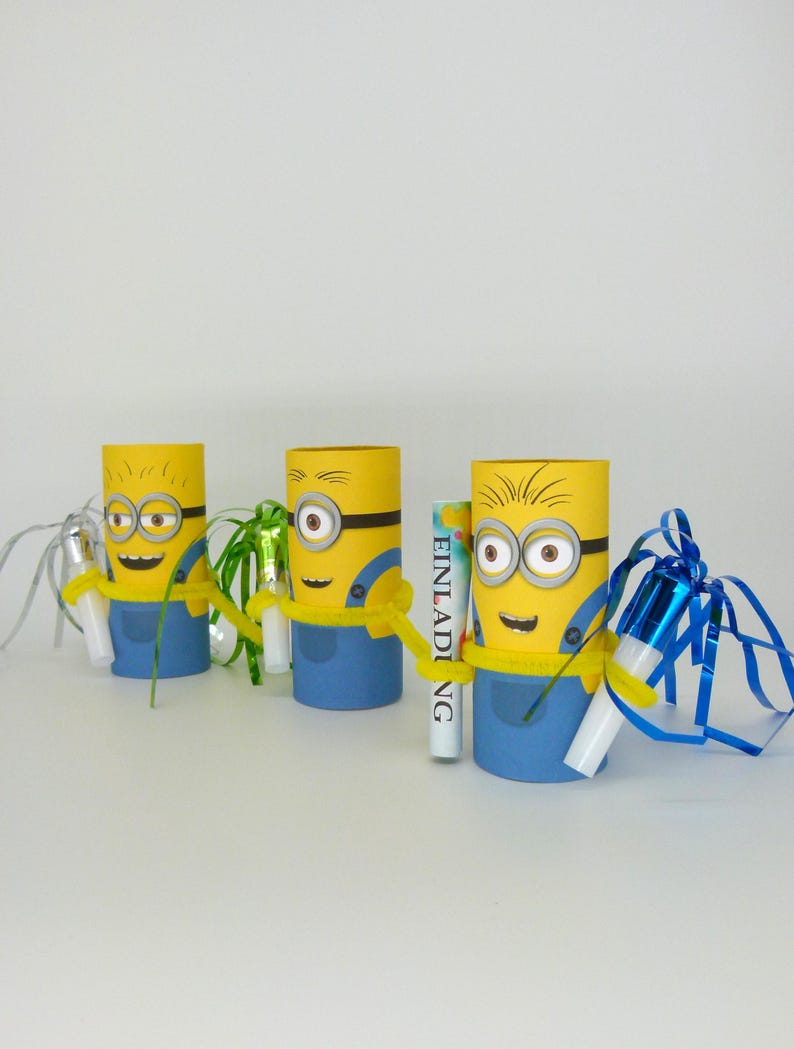 May include: Three yellow and blue Minion-themed cardboard tubes with party favors inside. Each tube has a different party favor, including a green and white party blower, a blue and white party blower, and a small white box with the word "Einladung" printed on it.