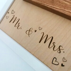 May include: A white framed wooden sign with the words "Mr. & Mrs." engraved in cursive script. The sign is decorated with small hearts.
