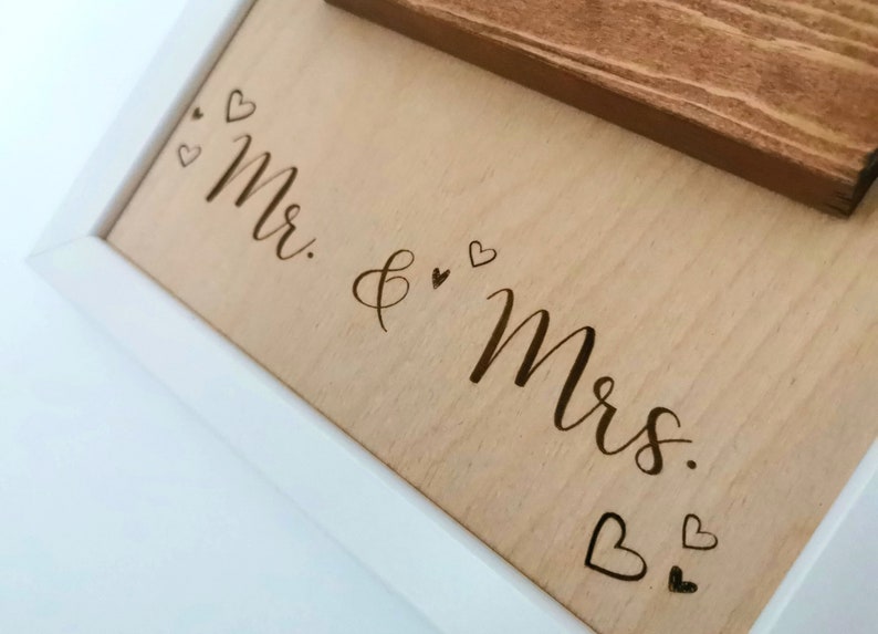 May include: A white framed wooden sign with the words "Mr. & Mrs." engraved in cursive script. The sign is decorated with small hearts.