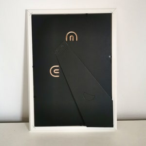 May include: White picture frame with a black backing and a fold-out easel stand. The easel stand has two slots for hanging the frame on a wall.