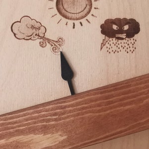 May include: A wooden weather vane with a black arrow pointing to the right. The vane has three weather symbols: a smiling sun, a cloud blowing wind, and a frowning cloud with rain and lightning.