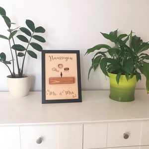 May include: A black framed wooden sign with the text "Hausseegen" and "Mr. & Mrs." in cursive. The sign has a small wooden plank with a sun, a bird, and a house drawn on it. The sign is sitting on a white dresser with two plants in pots.