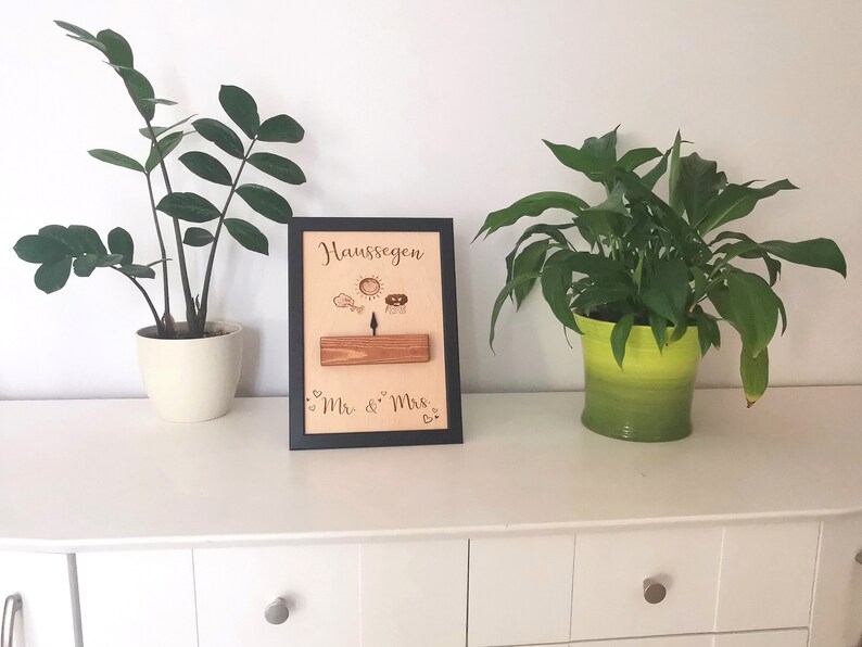 May include: A black framed wooden sign with the text "Hausseegen" and "Mr. & Mrs." in cursive. The sign has a small wooden plank with a sun, a bird, and a house drawn on it. The sign is sitting on a white dresser with two plants in pots.