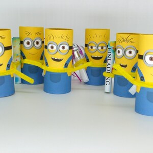 May include: Six yellow Minion character party favors made from cardboard toilet paper rolls. Each Minion has a blue jumpsuit, goggles, and a yellow pipe cleaner. The word "Einladung" is written on one of the favors.
