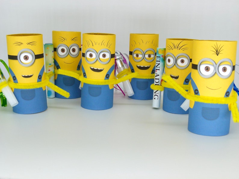 May include: Six yellow Minion character party favors made from cardboard toilet paper rolls. Each Minion has a blue jumpsuit, goggles, and a yellow pipe cleaner. The word "Einladung" is written on one of the favors.