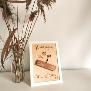 May include: A white framed wooden sign with the text "Haus Segen" and a small wooden swing with a sun, cloud, and raindrop design. The sign also has the text "Mr. & Mrs." with hearts.