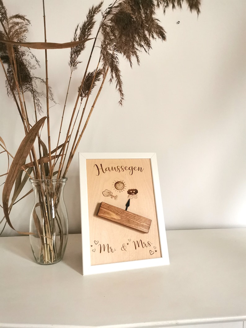 May include: A white framed wooden sign with the text "Haus Segen" and a small wooden swing with a sun, cloud, and raindrop design. The sign also has the text "Mr. & Mrs." with hearts.