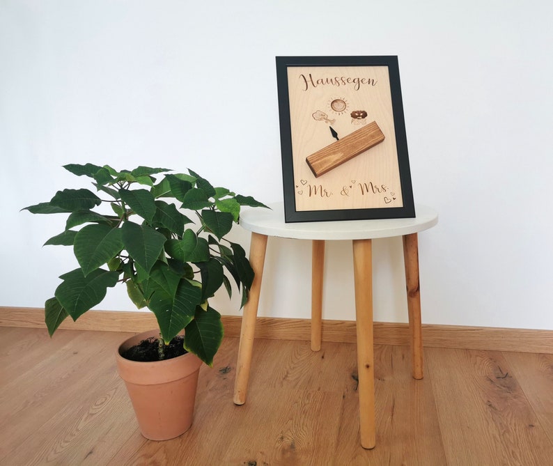 May include: A wooden plaque with the text "Haus Segen" and "Mr. & Mrs." in a black frame. The plaque has a small wooden piece attached to it with a sun, a ring, and a cloud carved into it. The plaque is sitting on a white table with a green plant in a terracotta pot in the foreground.