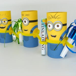 May include: Three yellow Minion-themed cardboard tubes decorated with blue overalls, googly eyes, and black goggles. Each tube has a different party favor inside, including a blue and silver party blower, a green streamer, and a white card with the word "Einladung" written on it.