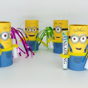 May include: Four yellow and blue Minion character party favor holders made from cardboard toilet paper rolls. Each holder has a yellow and blue Minion face with googly eyes and a yellow smile. Each holder has a different colored streamer attached to it. One holder has a white tube with the word "EINLADUNG" written on it.