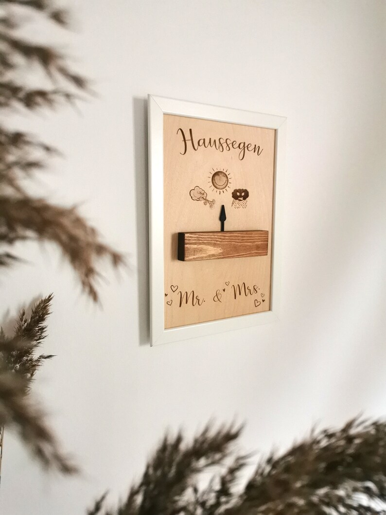 May include: A white framed wooden sign with the text "Haus Segen" and a sun, cloud, and arrow design. The sign also has the text "Mr. & Mrs." with hearts.