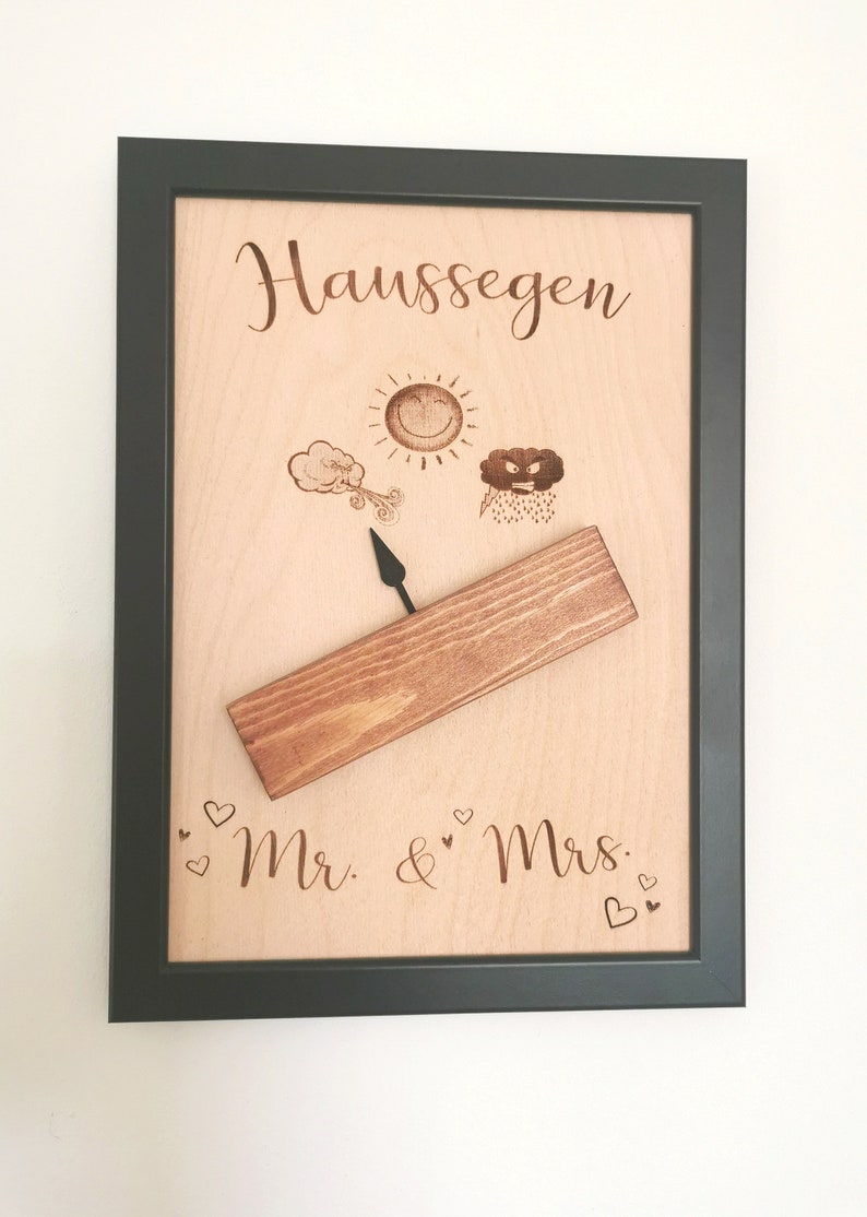 May include: A wooden plaque with a black frame, featuring the text "Haus Segen" and a weather vane with a sun, a cloud with wind, and a rain cloud. The plaque also includes the text "Mr. & Mrs." with hearts.