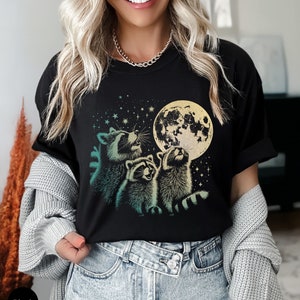 Op de afbeelding: Zwart T-shirt met een afbeelding van drie wasberen die naar een volle maan kijken met sterren op de achtergrond.
