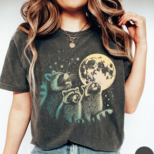 Op de afbeelding: Een donkergrijs T-shirt met een grafische print van drie wasberen die naar een gele maan met sterren op de achtergrond kijken.